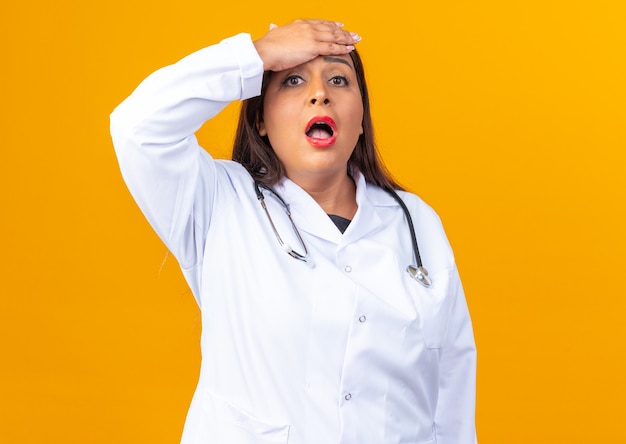 Medico donna di mezza età in camice bianco con stetoscopio stupito e sorpreso con la mano sulla fronte