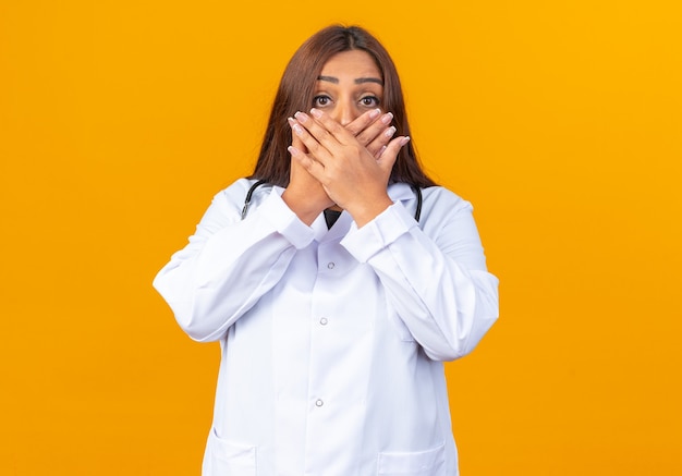 Middle age woman doctor in white coat with stethoscope being shocked covering mouth with hands standing over orange wall