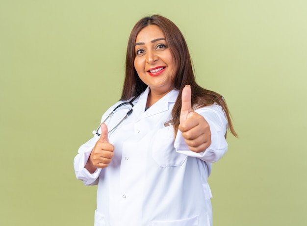 Женщина-врач среднего возраста в белом халате со стетоскопом счастлива и позитивно улыбается, весело показывает палец вверх, стоя над зеленой стеной