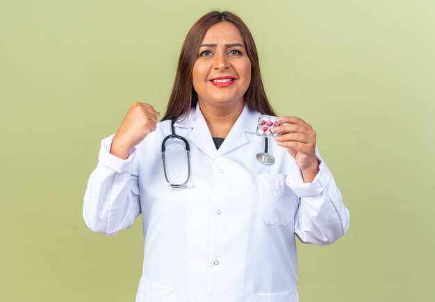 Middle age woman doctor in white coat with stethoscope holding blister with pills clenching fist smiling confident standing on green