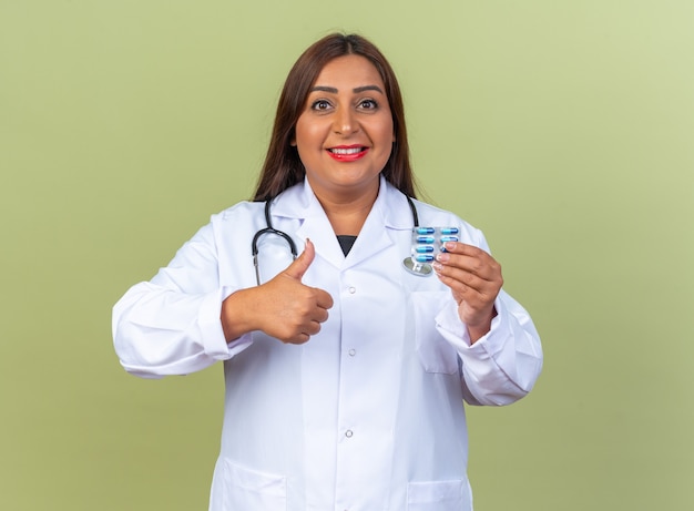 Middle age woman doctor in white coat with stethoscope holding blister with pills showing thumbs up smiling confident standing on green