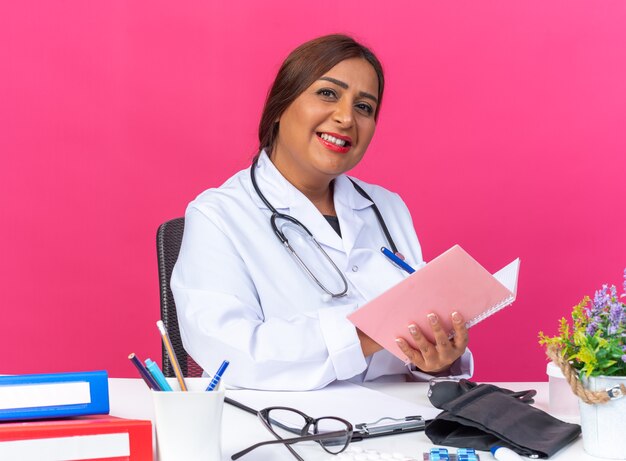 Женщина-врач среднего возраста в белом халате со стетоскопом, держащая блокнот, пишет что-то с улыбкой на счастливом лице, сидя за столом с офисными папками на розовом