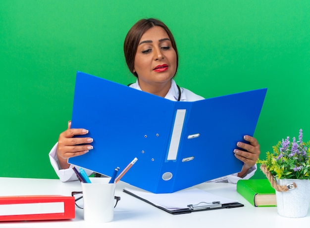 Middle age woman doctor in white coat with stethoscope holding open office folder looking at it intrigued sitting at the table on green