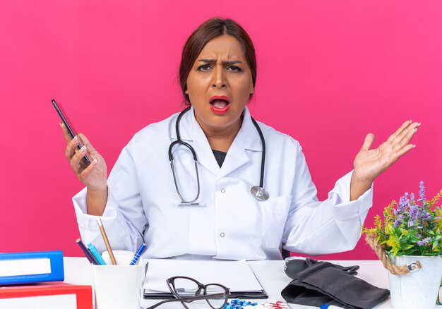 Женщина-врач среднего возраста в белом халате со стетоскопом, держащая смартфон в замешательстве, разводит руками в стороны, сидя за столом с офисными папками на розовом
