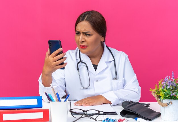 ピンクの背景の上のオフィスフォルダーとテーブルに座って不機嫌な深刻な顔でそれを見てスマートフォンを保持している聴診器と白衣を着た中年女性医師