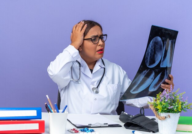 Middle age woman doctor in white coat with stethoscope holding x-ray looking at it with confuse expression with hand on her head sitting at the table over blue wall