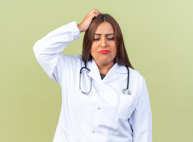 Женщина-врач среднего возраста в белом халате со стетоскопом выглядит озадаченно, почесывая голову, стоя над зеленой стеной