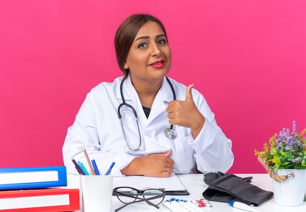 Женщина-врач среднего возраста в белом халате со стетоскопом, глядя, улыбаясь, уверенно показывает палец вверх, сидя за столом с офисными папками на розовом фоне