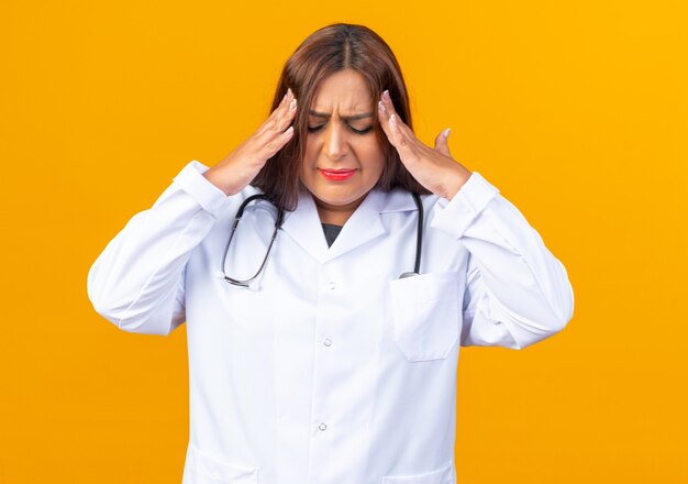 Middle age woman doctor in white coat with stethoscope looking unwell touching her head suffering from headache