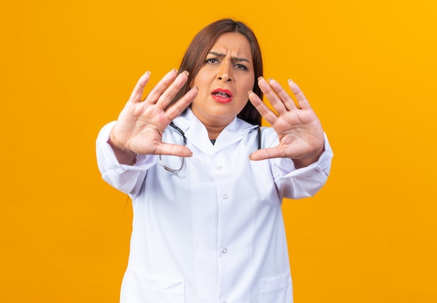 Middle age woman doctor in white coat with stethoscope looking with serious face making stop gesture with hands