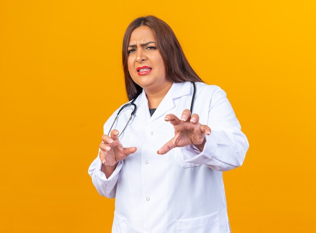 Middle age woman doctor in white coat with stethoscope looking worried and scared making defense gesture with hands