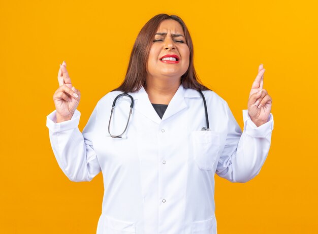 Middle age woman doctor in white coat with stethoscope making desirable wish with eyes closed with hope expression crossing fingers