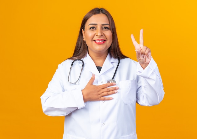 Medico della donna di mezza età in camice bianco con lo stetoscopio che fa gesto di giuramento che sembra sorridente fiducioso