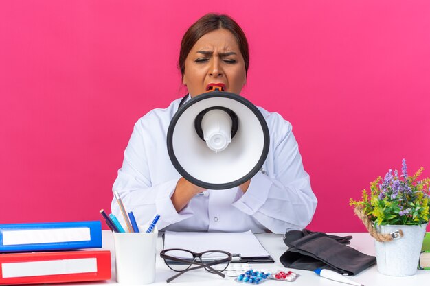 Middle age woman doctor in white coat with stethoscope shouting to megaphone being excited sitting at the table with office folders on pink