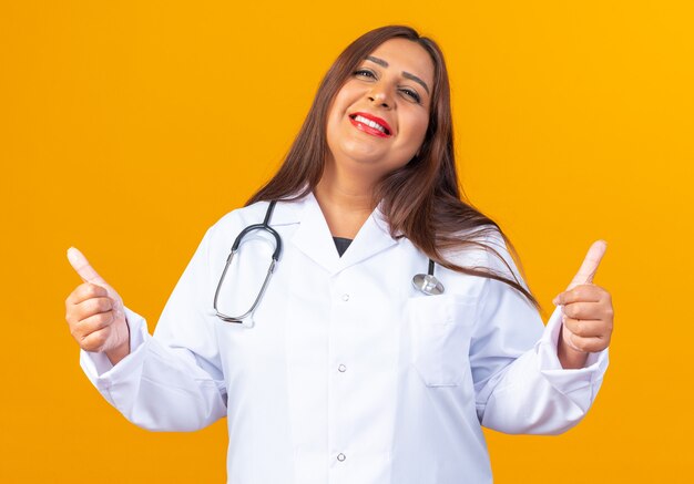 Женщина-врач среднего возраста в белом халате со стетоскопом, весело улыбаясь, счастливая и позитивная, показывает палец вверх