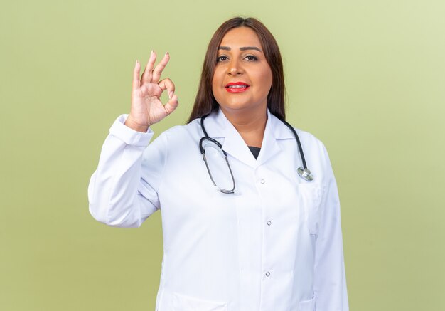 聴診器と白いコートを着た中年女性医師は、緑の上に立ってOKサインを自信を持って笑っています