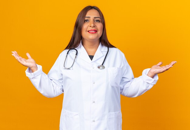 Женщина-врач среднего возраста в белом халате со стетоскопом, уверенно улыбаясь, разводит руками в стороны, стоя над оранжевой стеной
