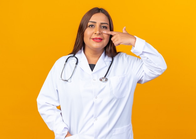 Женщина-врач среднего возраста в белом халате со стетоскопом улыбается, указывая указательным пальцем на нос, стоя над оранжевой стеной