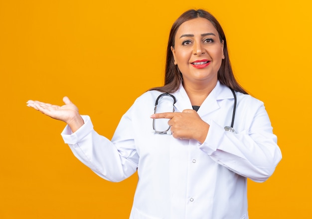 Medico della donna di mezza età in camice bianco con lo stetoscopio sorridente che indica con il dito indice al lato e che presenta qualcosa con il braccio che sta sopra la parete arancione