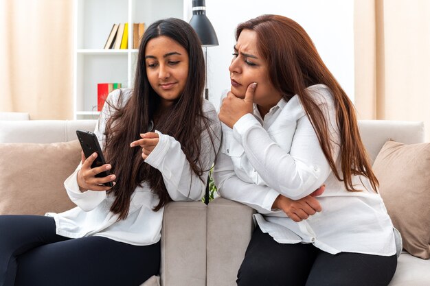 中年の女性と白いシャツと黒いズボンを着た若い娘が椅子に座り、スマートフォンを持つ娘と明るいリビングルームでスマートフォンが不機嫌になっているのを見ている母親