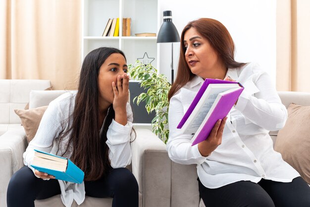 Middle age woman and her young daughter in white shirts and black pants sitting on the chairs with books daughter looking surprised at book in light living room