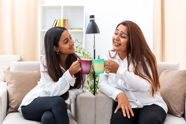 Middle age woman and her young daughter in white shirts and black pants sitting on the chairs with cups of hot tea happy and positive spending time together in light living room