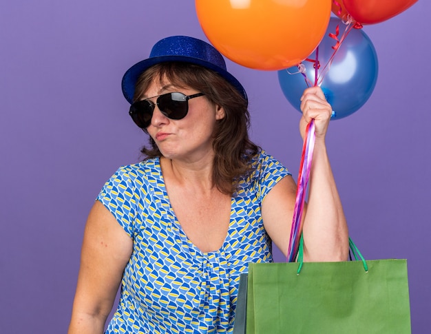 Женщина средних лет в шляпе и очках держит кучу разноцветных шаров и бумажных пакетов с подарками, выглядит смущенной и недовольной, празднует день рождения, стоя над фиолетовой стеной