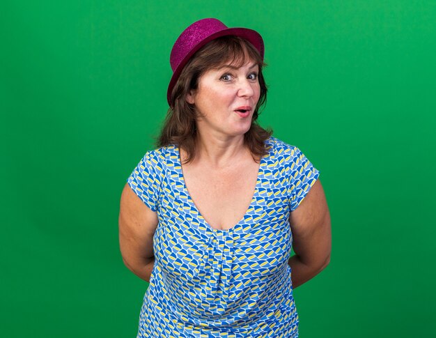 Middle age woman in party hat  happy and surprised celebrating birthday party standing over green wall