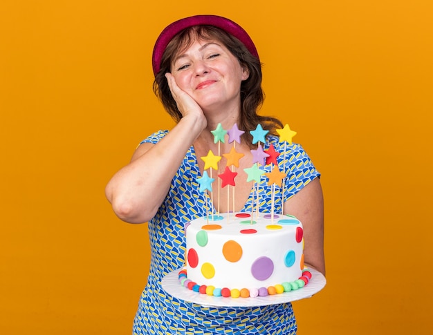 Donna di mezza età con cappello da festa che tiene in mano una torta di compleanno felice e positiva che sorride allegramente