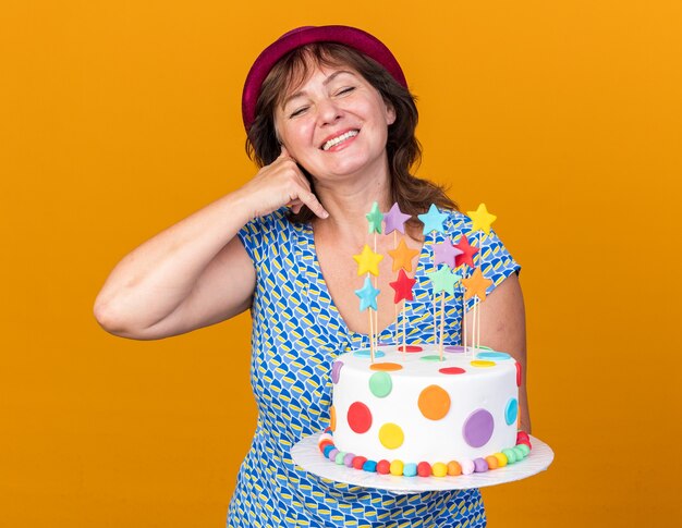 Middle age woman in party hat holding birthday cake smiling cheerfully happy and positive making call me gesture celebrating birthday party standing over orange wall