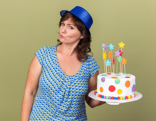 Middle age woman in party hat holding birthday cake  with skeptic expression celebrating birthday party standing over green wall