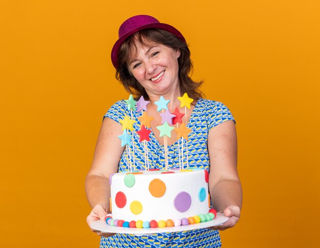 Middle age woman in party hat holding birthday cake  with smile on face happy and cheerful celebrating birthday party standing over orange wall