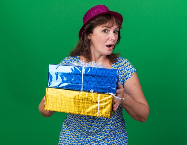 Middle age woman in party hat holding birthday gifts  surprised celebrating birthday party standing over green wall
