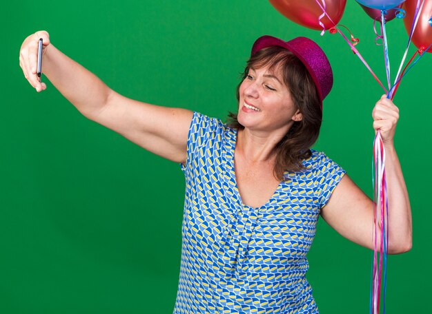 スマートフォンを使ってセルフィーをしているカラフルな風船を持ったパーティーハットの中年女性が、緑の壁の上に立って誕生日パーティーを祝う幸せで陽気な笑顔