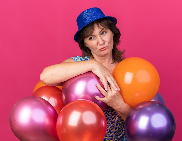 Middle age woman in party hat holding colorful balloons looking aside with skeptic expression