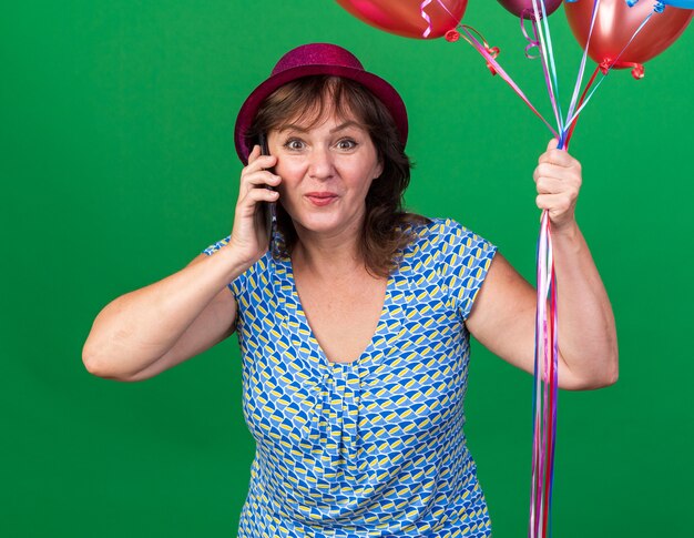 Middle age woman in party hat holding colorful balloons looking happy and surprised while talking on mobile phone celebrating birthday party standing over green wall