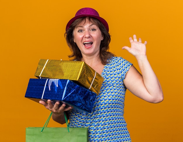 Middle age woman in party hat holding paper bag with birthday gifts  happy and cheerful smiling with arm raised