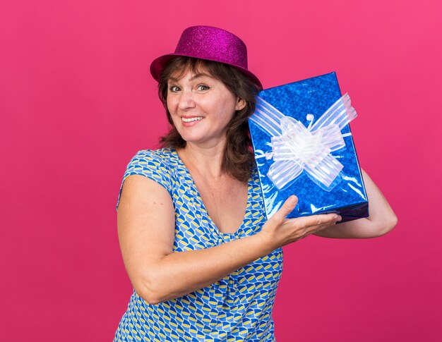 Middle age woman in party hat holding a present  happy and cheerful smiling broadly celebrating birthday party standing over pink wall