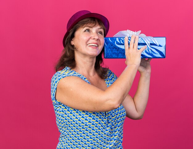 Middle age woman in party hat holding a present happy and cheerful smiling broadly