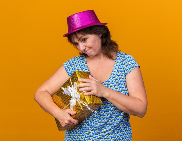 Middle age woman in party hat holding a present looking confused going to open box celebrating birthday party standing over orange wall