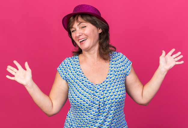 Middle age woman in party hat  smiling cheerfully with arms raised celebrating birthday party standing over pink wall