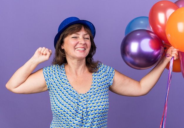 カラフルな風船の束が拳を握りしめているパーティーハットをかぶった中年女性は、紫色の壁の上に立って幸せで興奮して誕生日パーティーを祝う
