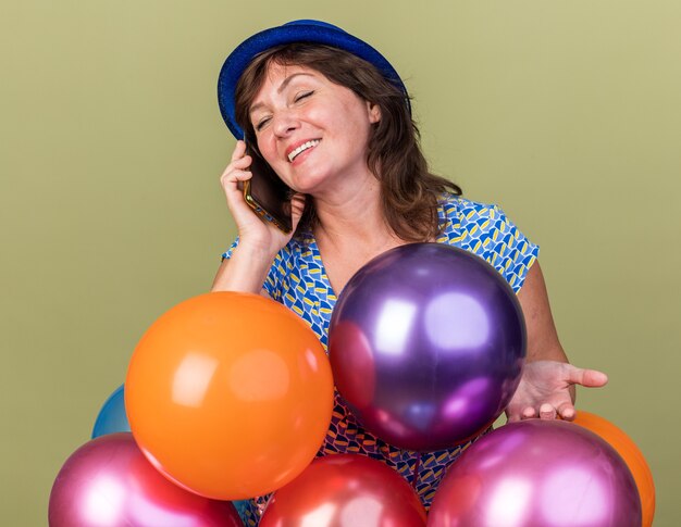 녹색 벽 위에 서있는 생일 파티를 축하하는 휴대 전화로 이야기하는 동안 유쾌하게 웃는 다채로운 풍선의 무리와 함께 파티 모자에 중년 여자