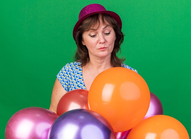 Middle age woman in party hat with colorful balloons looking down intrigued celebrating birthday party standing over green wall