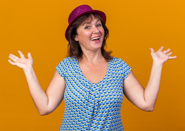 Middle age woman in party hat  with happy face smiling cheerfully raising arms