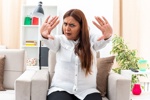Middle age woman in white shirt and black pants  with serious face making stop gesture with hands sitting on the chair in light living room