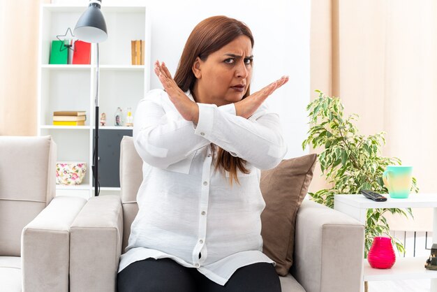 Middle age woman in white shirt and black pants  with serious frowning face making stop gesture crossing hands sitting on the chair in light living room