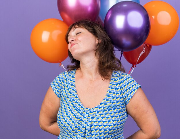 カラフルな風船の束を持つ中年の女性は、幸せで喜んで笑顔で、紫色の壁の上に立って誕生日パーティーを陽気に