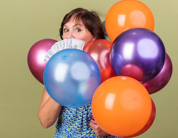 Middle age woman with bunch of colorful balloons holding cash  surprised celebrating birthday party standing over green wall