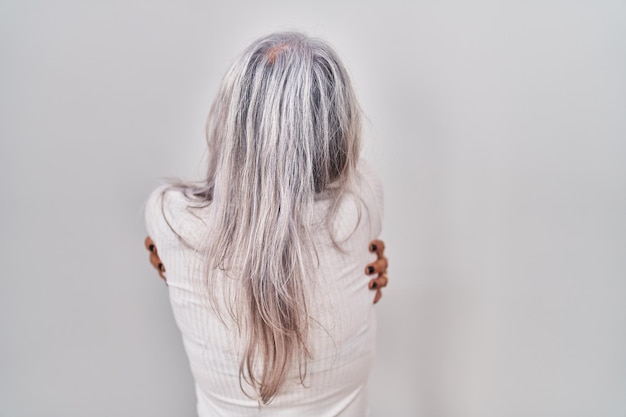 Middle age woman with grey hair standing over white background hugging oneself happy and positive from backwards self love and self care